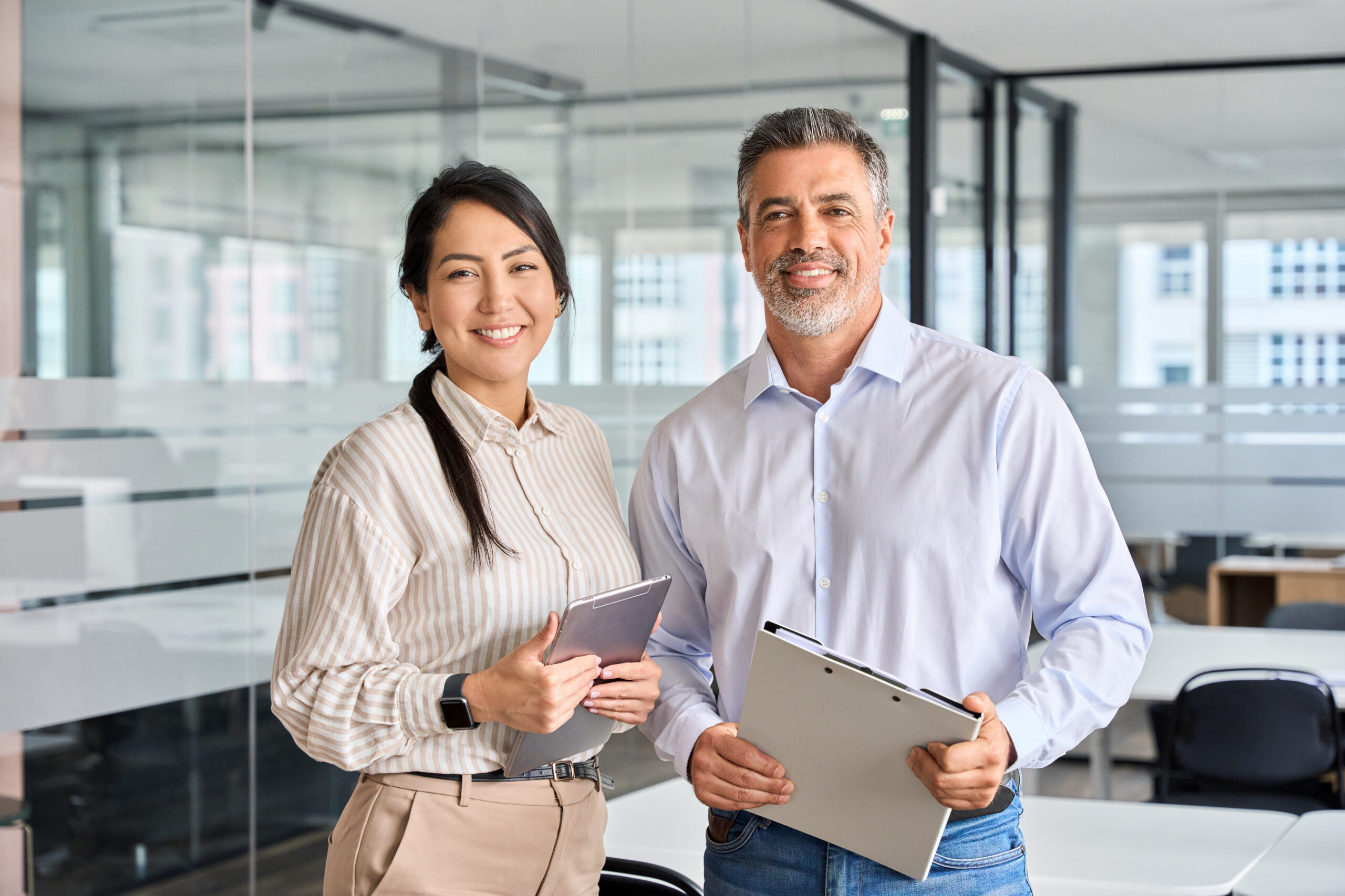 Happy confident professional mature Latin business man and Asian business woman corporate leaders managers standing in office, two diverse colleagues executives team posing together, portrait.